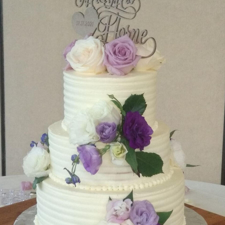 Three-tier buttercream wedding cake with purple and white roses and a monogram topper