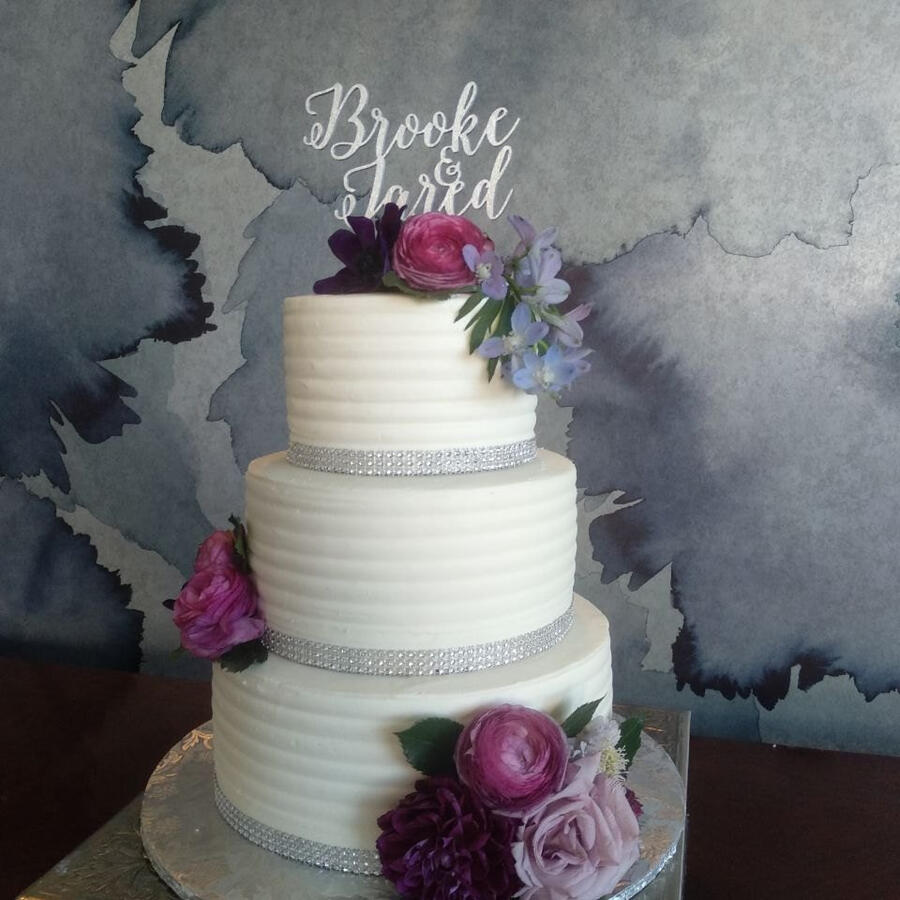 Tiered white wedding cake with purple flowers and a script name topper Tiered white wedding cake with purple flowers and a script name topper