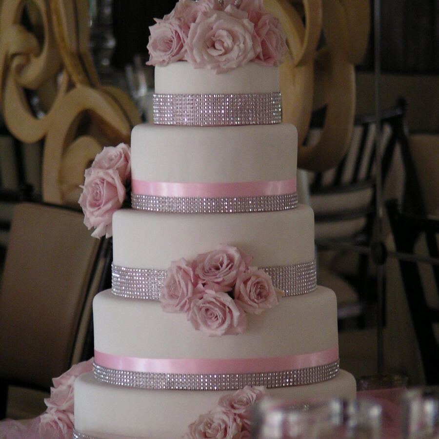 Four-tier white wedding cake decorated with pink roses and pink ribbon in an ornate ballroom