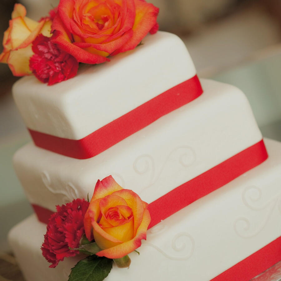 Square tiered white cake with red ribbon and orange and pink roses